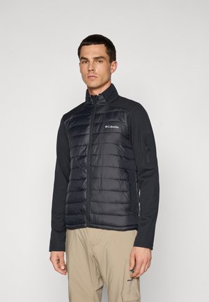 Mann trägt eine schwarze Columbia Reißverschluss-Puffjacke mit langen Ärmeln und beigen Cargo-Hosen, steht vor einem einfarbigen Hintergrund.