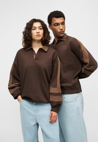 Καφέ πουλόβερ με γιακά και μακριά μανίκια, με τονικά πάνελ ως διακοσμητικά στοιχεία. Συνδυασμένο με ανοιχτόχρωμα, χαλαρά τζιν παντελόνια.