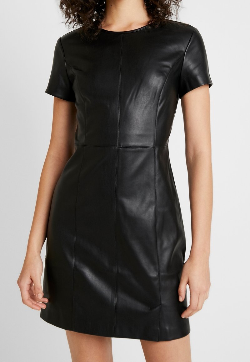 Femme portant une robe en cuir noire à manches courtes avec des détails de couture, une taille cintrée et un col rond, sur un fond uni.