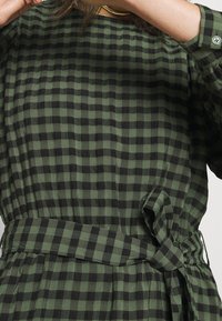 Vestido a cuadros en verde y negro, confeccionado con una tela ligera, que presenta un cinturón atado en la cintura, mangas largas y un diseño de corte holgado.
