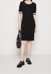 Femme portant une robe noire mi-longue avec des boutons latéraux, des poignets de manches rayés, tenant un sac à main noir texturé et des mocassins noirs avec des chaînes dorées.