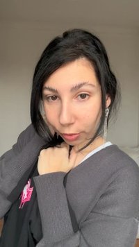 Νέα γυναίκα με σκούρα μαλλιά, φορόντας γκρι πουλόβερ και μακρύ ασημένιο σκουλαρίκι, ακουμπώντας το χέρι της κοντά στο πρόσωπο, κοιτάζοντας κατευθείαν στην κάμερα.