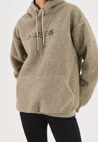 Tøjdunkel beige hættetrøje med en tekstureret yderside, snøre i hætten og frontlomme. Har sort skrift på brystet. Ribbet kant.