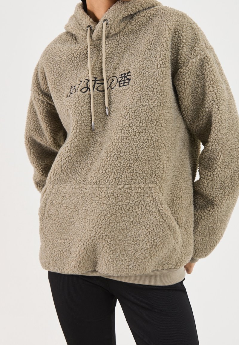 Tøjdunkel beige hættetrøje med en tekstureret yderside, snøre i hætten og frontlomme. Har sort skrift på brystet. Ribbet kant.