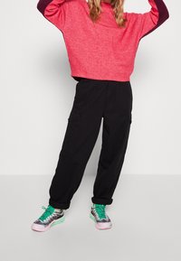 Roter und lila weit geschnittener, gekürzter Pullover, kombiniert mit schwarzen Cargo-Hosen und mehrfarbigen Sneakers mit pinken, grünen und grauen Akzenten.