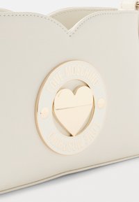 Dettaglio di una borsa in pelle crema con emblema a forma di cuore dorato incorniciato da una placca circolare con il testo "Love Moschino".