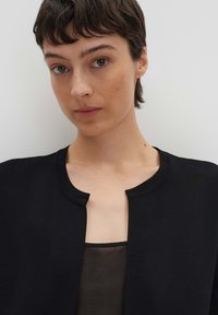 Cardigan en tricot noir avec un col rond, porté par-dessus un top sombre et transparent. Le tissu a une finition texturée, et le design est ouvert à l'avant.