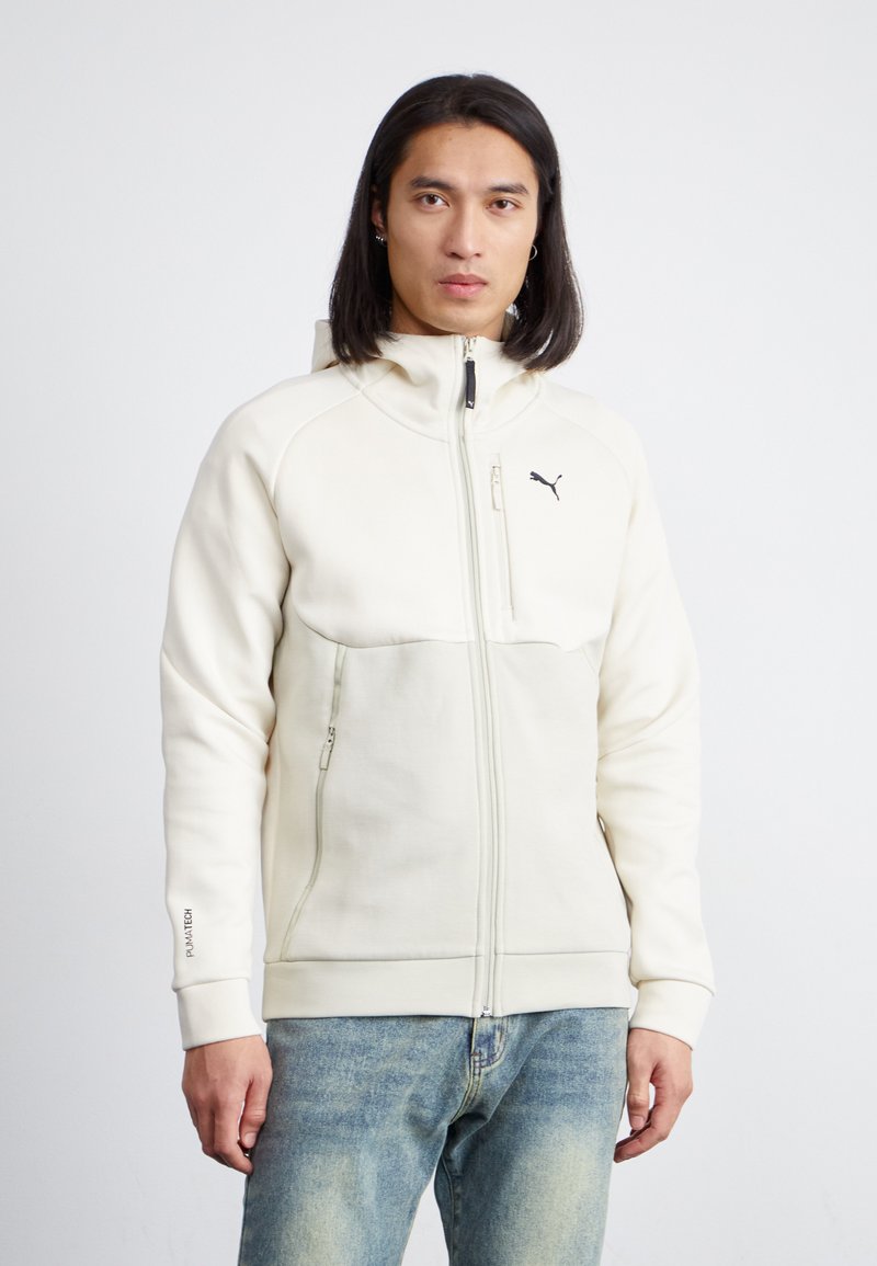 Hvit zip-up hettegenser med høy krage, kontrastpaneler, frontlommer, og en svart logo på brystet. Brukt med lyseblå jeans.
