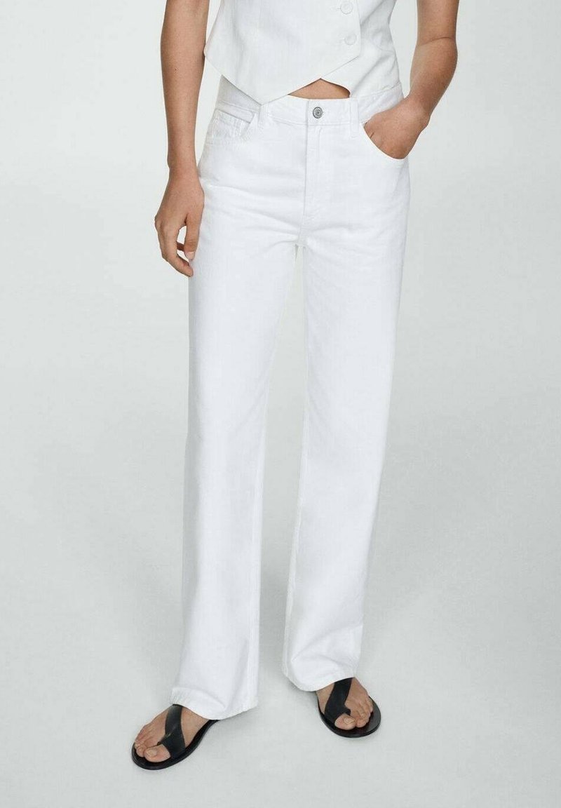Personne portant un jean blanc taille haute à jambe droite, un haut blanc sans manches avec un ourlet en encoches, et des sandales noires à brides croisées, debout avec une main dans la poche.