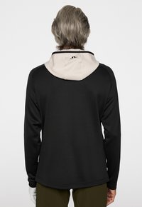 Hoodie noir à manches longues avec une capuche beige contrastante. Présente un logo au dos et un tissu lisse et texturé.