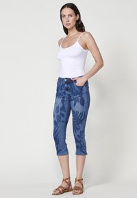 Jean capri en denim bleu avec un motif floral, taille mi-haute et design à cinq poches, associé à un débardeur blanc et des sandales brunes à lanières.