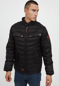 Black padded jacket with a high collar, two zippered chest pockets, and contrasting brown leather accents. Textured fabric with visible stitching.