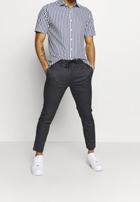 Hombre que lleva zapatillas blancas, pantalones negros de rayas con cordón y una camisa de botones de manga corta a rayas verticales en azul y blanco.