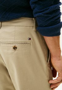 Pantaloni beige con tessuto testurizzato. Presentano una tasca posteriore con bottone e un piccolo patch logo multicolore vicino alla tasca.