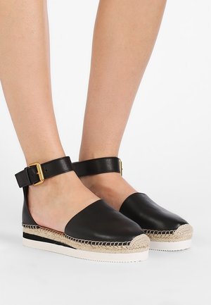 Fødder iført sorte læder espadrills med flade såler, ankelstropper og guldlås på hvid baggrund.