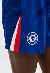 Blå Chelsea Football Club-shorts med røde og hvite sidestriper, båret på en persons lår.