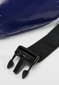 Sac bleu brillant avec une sangle noire épaisse et une boucle en plastique, présentant des crêtes distinctes sur la sangle et une surface lisse et brillante.