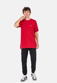 Camiseta de algodón roja de manga corta y cuello redondo. Combinada con pantalones negros y zapatillas en blanco y negro. Detalle de pequeño logo en el pecho.