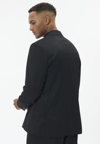 Juodas pritaikytas švarkas, pagamintas iš tekstūruotos medžiagos. Vieno mygtuko modelis, šiek tiek iškirptais atvartais ir sagomis rankogaliuose. Minimalistinis stilius ir švarios linijos.