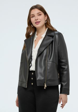 Nainen pukeutunut mustaan nahkatakkiin valkoisen napillisen paidan ja korkeavyötäröisten mustien housujen kanssa, joissa on kullanväriset napit, yksinkertaista taustaa vasten.