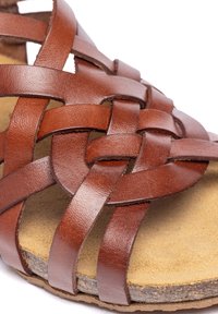 Sandalia de cuero marrón con diseño de tiras trenzadas, suela plana y textura del plantillaje en beige. Presenta un patrón de cruz y un acabado natural.