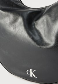 Μαύρη δερμάτινη τσάντα με ασημένιο φερμουάρ και λευκό λογότυπο "cK" στη κάτω δεξιά γωνία, σε απλό φόντο.