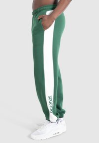 Person iført grønne sweatpants med hvide sidestriber og "SMILODOX" tekst, kombineret med hvide sneakers, stående mod en ensfarvet baggrund.