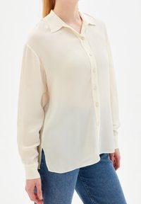 Femme portant un chemisier crémeux à manches longues et larges avec des boutons et un jean bleu, se tenant devant un fond blanc uni.