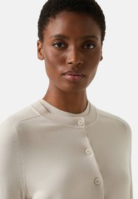 Beige cardigan made of soft knit fabric, featuring a round neck, four matching buttons, and subtle stitching along the shoulders.