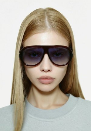 Femme aux longs cheveux blonds et raides portant de grandes lunettes de soleil dégradées et surdimensionnées, vêtue d'un haut gris clair à col rond, face à l'avant sur un fond blanc.