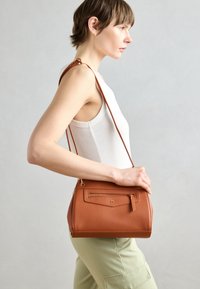 Bolso de hombro de cuero marrón con una forma estructurada, bolsillo frontal con cremallera y correa ajustable, que presenta una textura granulada y un diseño minimalista.