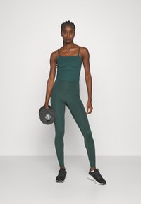Débardeur côtelé vert foncé à fines bretelles, associé à un legging assorti taille haute. Le modèle tient un haltère noir et porte des baskets noires.