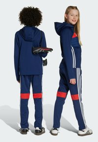 Chándal azul marino con detalles rojos y rayas blancas. Presenta una capucha y un corte atlético. Botas de fútbol negras y rojas sostenidas en una mano.