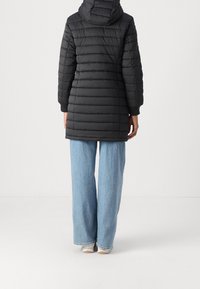 Chaqueta de abrigo negra con costuras horizontales, capucha y puños acanalados, llevada sobre unos vaqueros de pierna ancha de color azul claro.