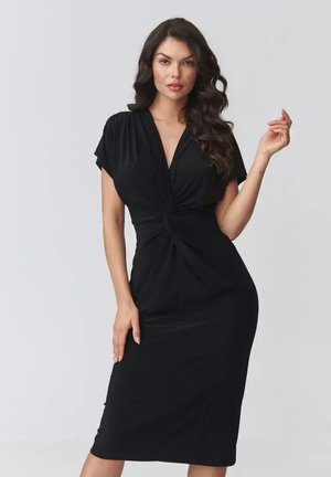 Kobieta o długich kręconych włosach, ubrana w dopasowaną czarną sukienkę do kolan z skręconym przodem, stojąca na tle jednolitego jasnego tła.