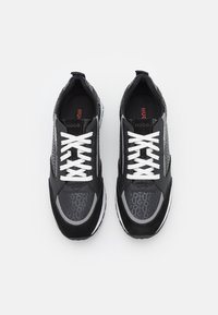 Chaussures de sport noires avec des lacets blancs, présentant un mélange de matériaux lisses et texturés, un motif de logo sur les côtés et une semelle blanche.