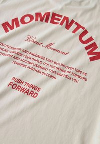 Biała koszulka z odważnym czerwonym napisem. "MOMENTUM" łukiem u góry, z dodatkowymi motywacyjnymi frazami poniżej w różnych rozmiarach i stylach.