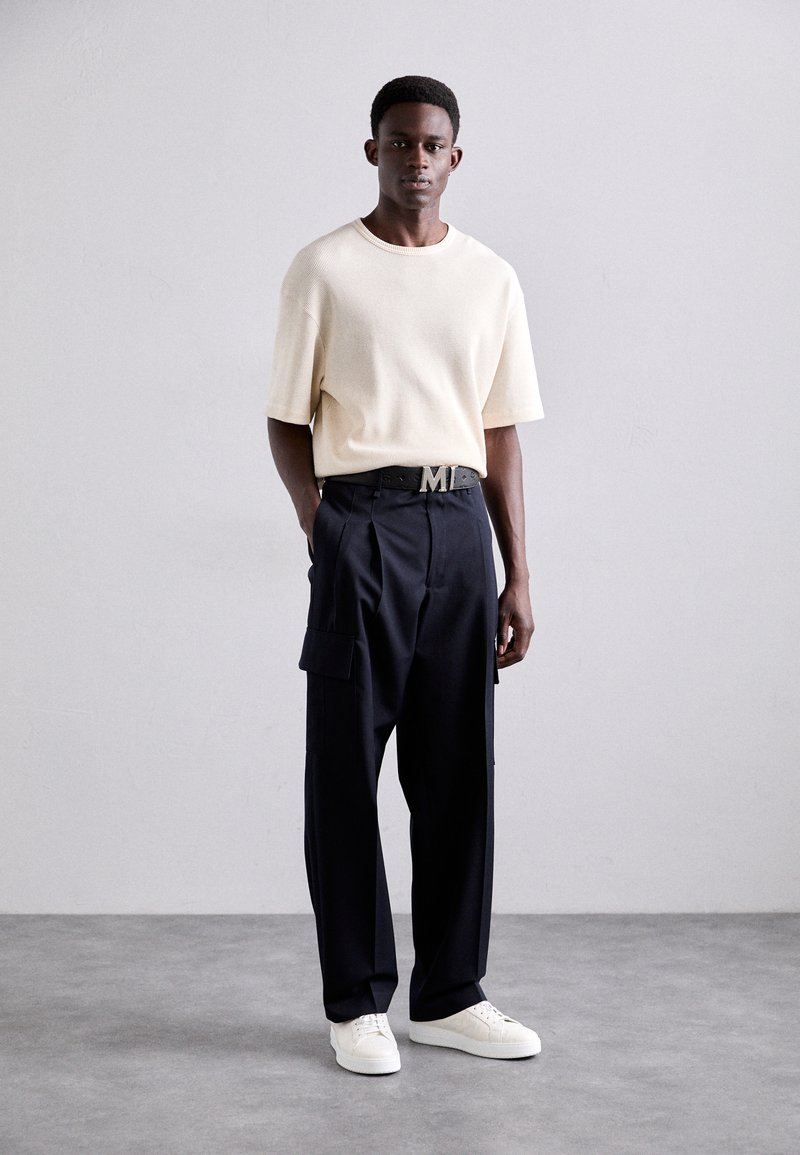 Cream short-sleeve textured knit shirt, black cargo trousers with side pockets, and white sneakers. Black belt with metal logo accent.