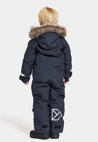 Marinblå overall med en pälsfodrad huva, justerbara ärmslut och en vit logotyp på vänster ben. Vattentätt material, vadderad design.