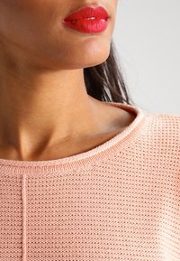 Ljusrosa stickad tröja med rund halsringning. Tyget har ett texturerat mönster och det finns synliga sömmar längs kanterna.