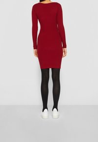 Rotes geripptes Strickkleid mit langen Ärmeln, figurbetonte Silhouette, getragen mit schwarzen Strumpfhosen und weißen Sneakers, vor einem schlichten Hintergrund präsentiert.