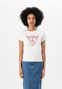 T-shirt bianco in cotone con logo triangolare rosso di Guess e dettagli in cristallo sul fronte, abbinato a una gonna di jeans.