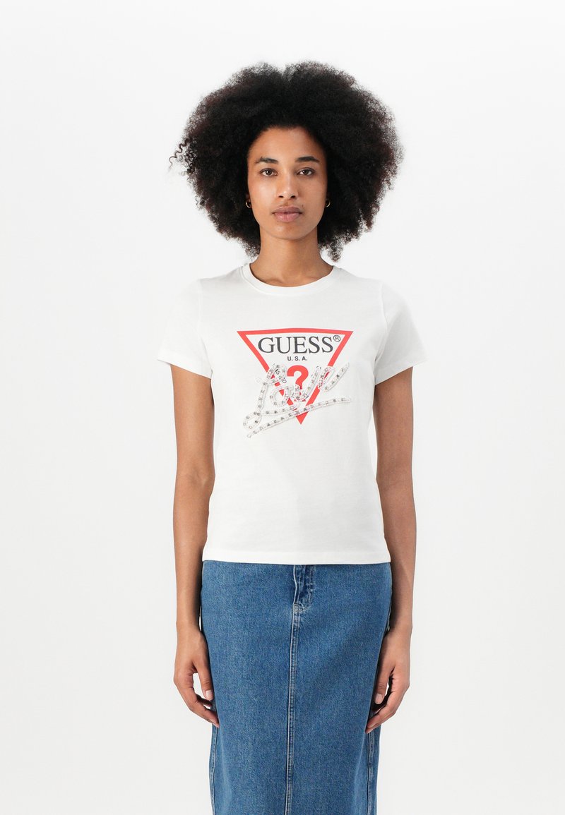 T-shirt bianco in cotone con logo triangolare rosso di Guess e dettagli in cristallo sul fronte, abbinato a una gonna di jeans.