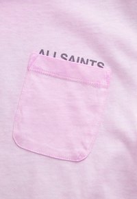 Gaiši rozā T-krekls ar kvadrātu kabatu, kuras krāsa ir saskaņota, virs kuras melnā krāsā ir drukāts teksts "ALL SAINTS", ar mīkstu auduma tekstūru.