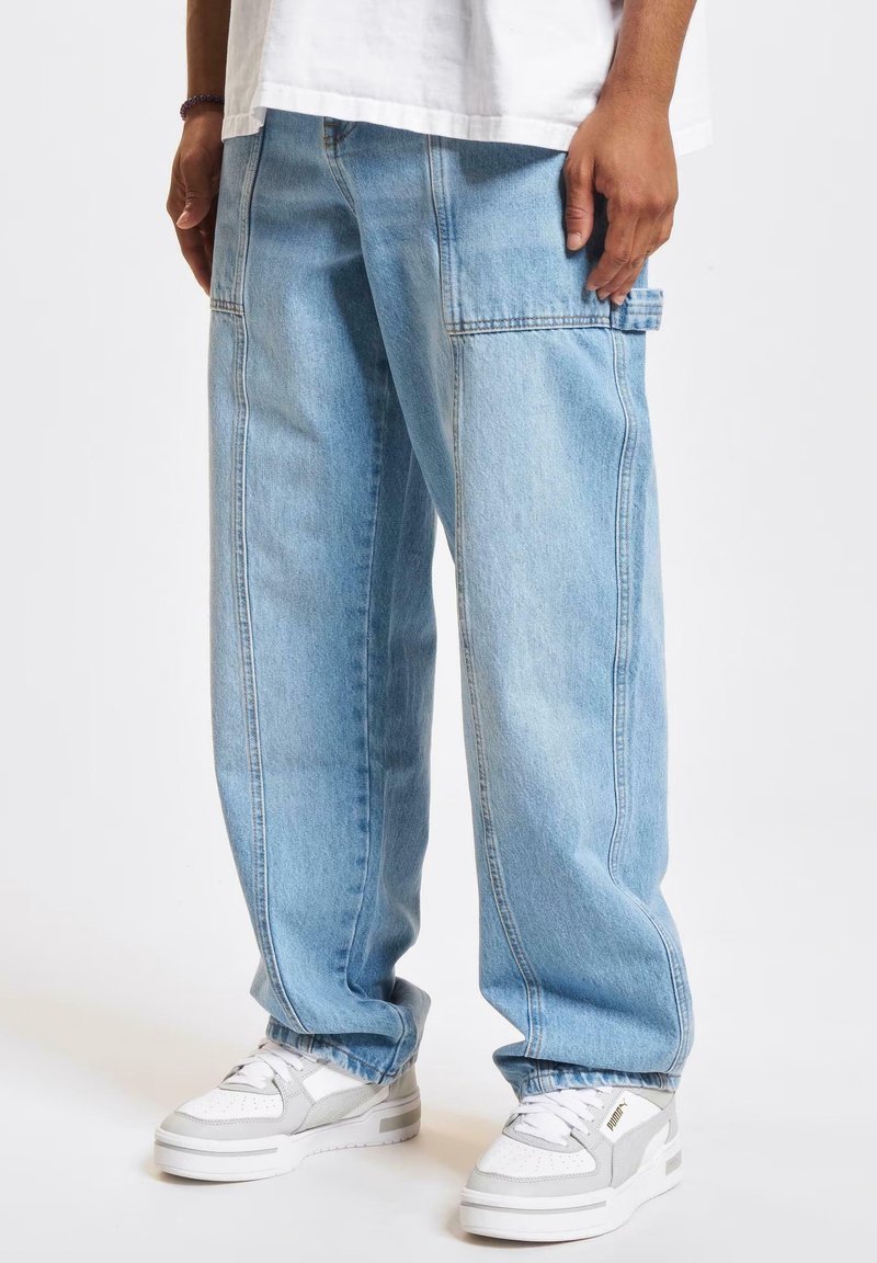 Homme portant un jean large bleu clair et des baskets blanches Puma, debout devant un fond blanc uni.