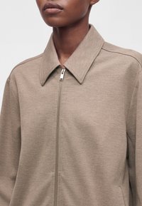 Gros plan d'une personne portant une veste beige à fermeture éclair avec un col pointu sur un fond clair uni.