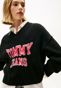 Schwarzer Strickpullover mit V-Ausschnitt, der das auffällige rote "TOMMY JEANS"-Logo zeigt. Hat gerippte Bündchen und Saum und ist über ein weißes Hemd mit Kragen getragen.