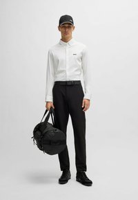 White long-sleeve button-up shirt with 'BOSS' logo, black belt and trousers, black sneakers, and a black duffel bag in hand.