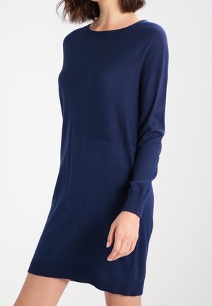 Woman wearing a long-sleeve, navy blue, knee-length sweater dress with a crew neckline, standing against a plain white background.