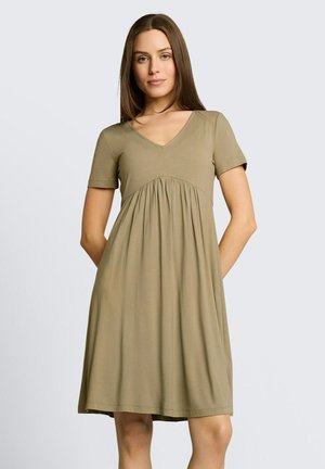 Vrouw met stijlbruin haar, gekleed in een olijfgroene, knielange jurk met korte mouwen, V-hals en een geplooide taille, staand tegen een effen achtergrond.